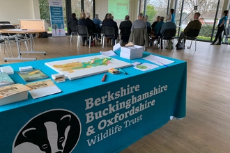 In the foreground is a table covered in a cloth with the BBOWT logo. On the table are flyers, a map, and other items. In the background a group of people are sat watching a presentation