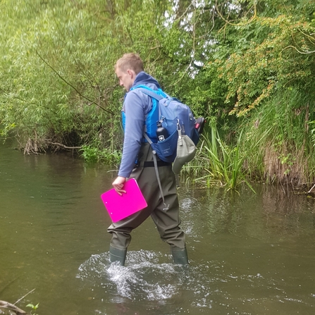 Person in waders walking through a shallow stream carrying a rucksack and clipboard