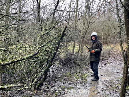 Person wearing full waterproofs and hard hat standing in mud in front of cut branches