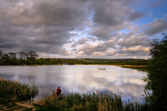 A man sits by the side of a lake looking over the water with moody clouds above him