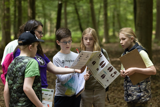 Group of people in a woodland looking at a mammal tracks and signs fold out booklet. There is one Wildlife Trust leader and four teenagers looking at the booklet.