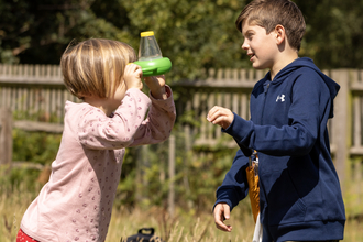 Two children in meadow. Child on left is looking into a two-way viewer bug pot and is wearing a pink long sleeved t-shirt, red spotty leggings and has short blonde hair. The child on the right is taller and is wearing a blue long-sleeved hoodie and is looking to also look into the bug viewer.  