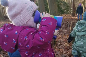 In woodland. Toddler wearing purple snowsuit and pink hat with bobbly ears is using children's binoculars and looking to the right. 