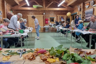 An image of a converted barn, full of people working at tables in the background. In the foreground is a table with a selection of fresh and dried leaves.