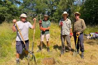 A group of four men standing in a woodland clearing with various tools.