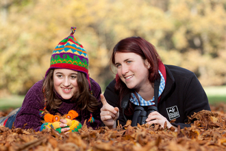 A Wildlife Trust member of staff pointing to something of interest to a happy young girl, dressed for autumn and surrounded by leaves