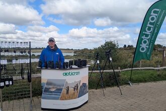 Man from Opticron at desk with binoculars and flag with view over College Lake 