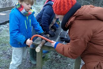Outside in winter, a boy in a blue jacket sawing a log whilst being guided by someone in a brown coat and red hat 