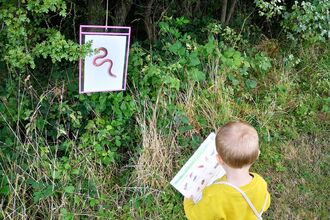 a toddler in a yellow jumper with a red bag, looking at a sheet amongst grass and shrubbery