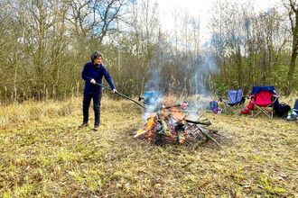 A person prodding a large stick into a bonfire in a winter woodland clearing. 
