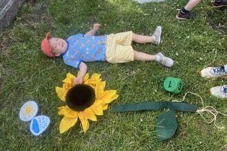 Image of a child lying down face-up on the grass, alongside a hand-made sculpture of a sunflower lying flat on the ground.