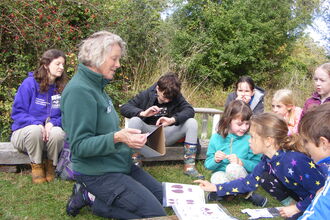 Image of a woman showing a group of kids how to identify animal tracks using identification posters. The children are sat outside in the grass looking interested.