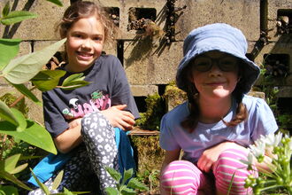 Portrait of two children outside amongst a wildlife garden. The sun is shining, they are smiling, and flowers and leaves are visible around them.
