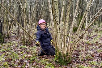 A person kneeling at the base of a tree in winter clothing, holding a cut branch in their hand.