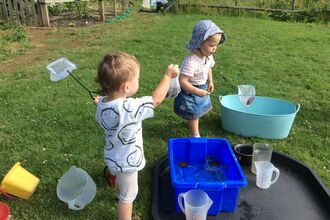 Image of two young children playing with pond-dipping equipment. They have nets in their hands and are looking at pond creatures in containers