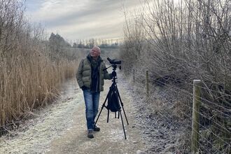 Man stands with scope and tripod on college lake reedbed path