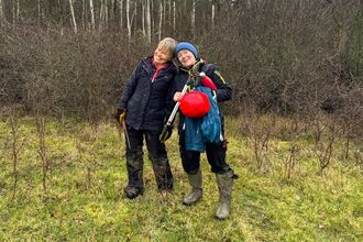 Two people in coats and boots holidng tree-cutting implements, smiling at the camera with winter hedges in the background.