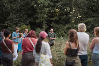 Image of a group of people in long grass, stood around a young man who is talking about nature. 