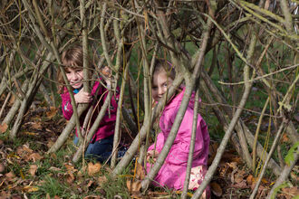 Image of two children playing on the ground of a Willow tunnel. They are partly obscured by the Willow whips, but their faces can be seen smiling.