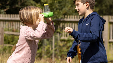 Two children in meadow. Child on left is looking into a two-way viewer bug pot and is wearing a pink long sleeved t-shirt, red spotty leggings and has short blonde hair. The child on the right is taller and is wearing a blue long-sleeved hoodie and is looking to also look into the bug viewer.