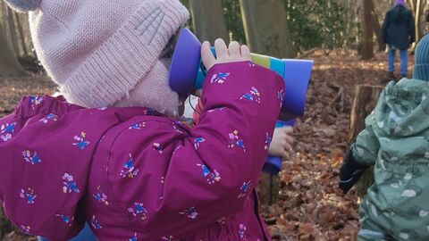 In woodland. Toddler wearing purple snowsuit and pink hat with bobbly ears is using children's binoculars and looking to the right. 