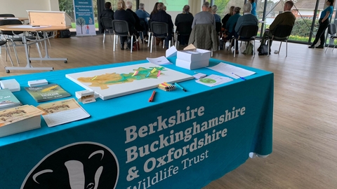 In the foreground is a table covered in a cloth with the BBOWT logo. On the table are flyers, a map, and other items. In the background a group of people are sat watching a presentation