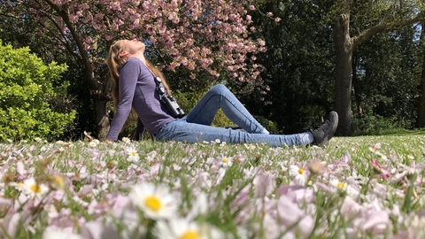 A woman sitting on the grass in a park, surrounded by flowers and trees, her face turned towards the sun