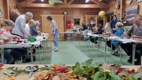 An image of a converted barn, full of people working at tables in the background. In the foreground is a table with a selection of fresh and dried leaves.