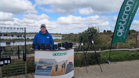 Man from Opticron at desk with binoculars and flag with view over College Lake 