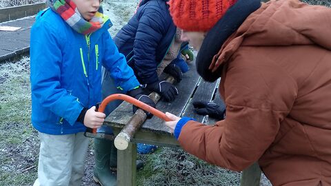 Outside in winter, a boy in a blue jacket sawing a log whilst being guided by someone in a brown coat and red hat 