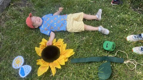 Image of a child lying down face-up on the grass, alongside a hand-made sculpture of a sunflower lying flat on the ground.