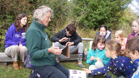 Image of a woman showing a group of kids how to identify animal tracks using identification posters. The children are sat outside in the grass looking interested.
