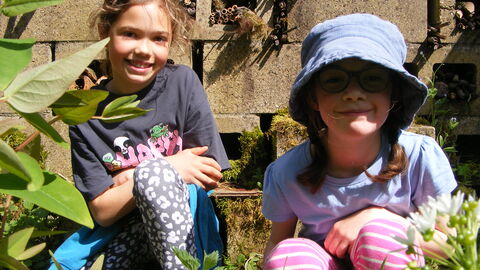 Portrait of two children outside amongst a wildlife garden. The sun is shining, they are smiling, and flowers and leaves are visible around them.