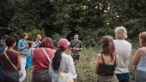 Image of a group of people in long grass, stood around a young man who is talking about nature. 