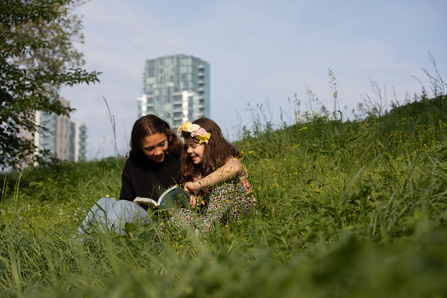 Woman and girl sitting in long grass reading a book in front of a tower block