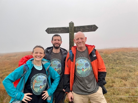 A woman and two men stood in front of a sign post on the Pennine way. The weather is misty.