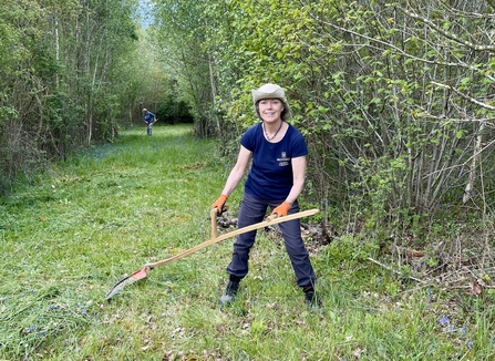 Woman scything grass on a grassy track surrounded by trees and shrubs