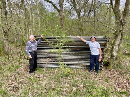 Two volunteers standing by a large pile of old Heras fencing in a woodland