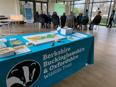 In the foreground is a table covered in a cloth with the BBOWT logo. On the table are flyers, a map, and other items. In the background a group of people are sat watching a presentation