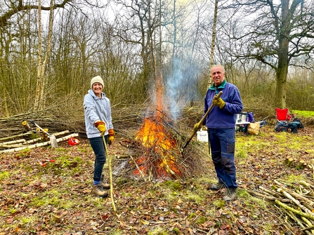A woman and man stood either side of a bonfire, with branches on the ground behind them.
