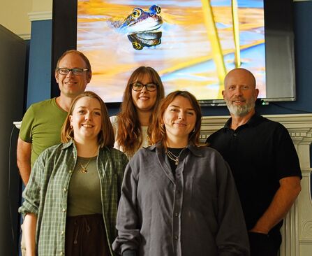 5 people smiling at the camera, stood in front of a digital screen displaying the frog in a pool which won the photo competition.