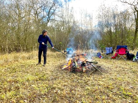 A person prodding a large stick into a bonfire in a winter woodland clearing. 