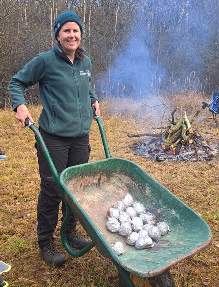 Blog author Charlotte Karmali standing with a large wheelbarrow filled with potatoes wrapped in foil. The bonfire can be seen in the background.