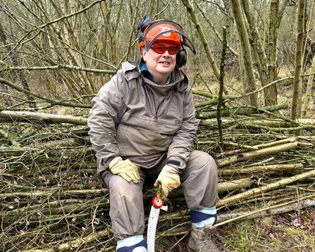 A person sat on a hedge of cut branches, with saw in hand.