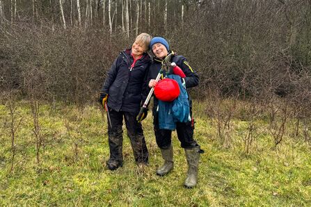 Two people in coats and boots holidng tree-cutting implements, smiling at the camera with winter hedges in the background.