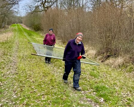 Two volunteers carrying a metal fence panel along a woodland ride.