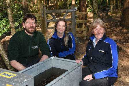Three people crouched next to a water tank in a woodland
