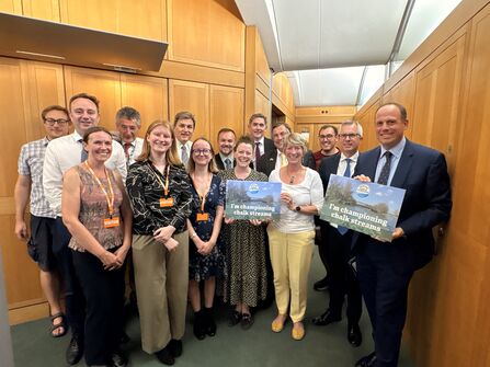 A group of people dressed smartly in a hallway, smiling at the camera. Two are holding signs which read: I am championing chalk streams